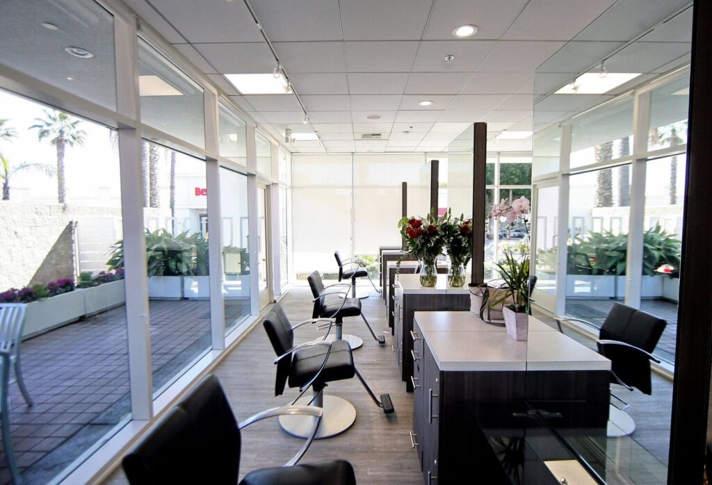 Hair salon interior with styling chairs, large mirrors, and floral arrangements near windows.